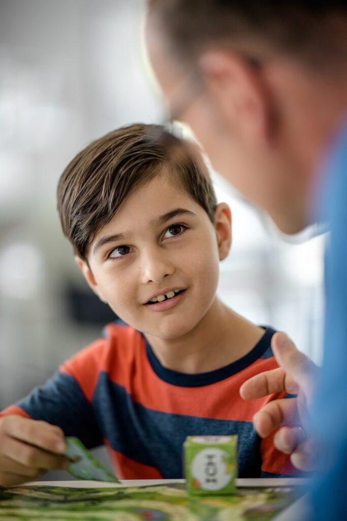 Foto von einem Jungen, der vor einem Spiel an einem Tisch sitzt und eine erwachsene Person anblickt, die nur im Anschnitt und unscharf zu sehen ist